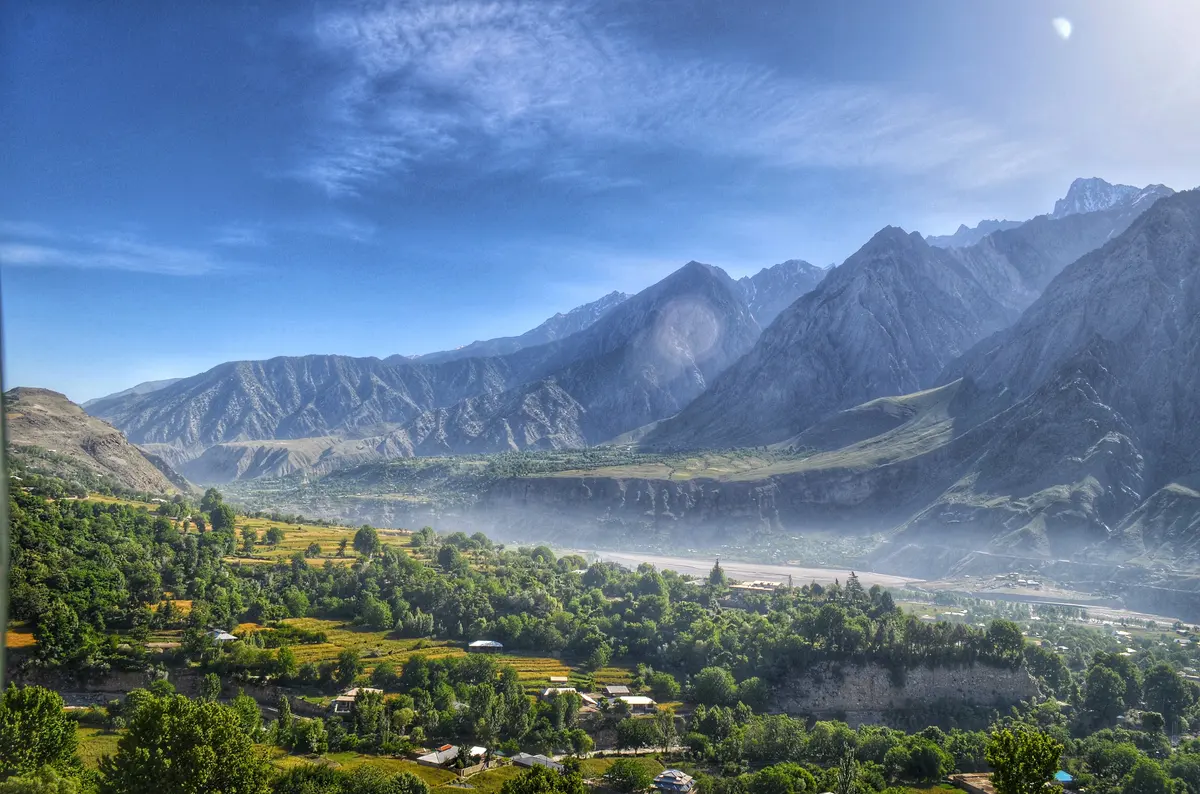 Terraced fields and orchards across a broad Chitral valley, with hazy Hindu Kush ridges beyond