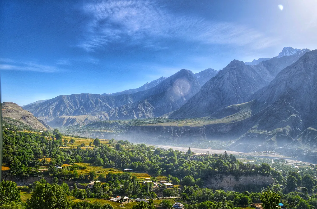 Terraced fields and orchards across a broad Chitral valley, with hazy Hindu Kush ridges beyond